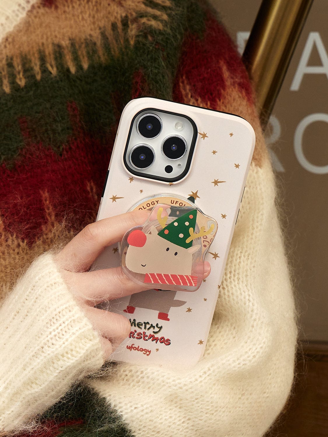 A hand holding an iPhone 16 Pro Max with a Rudolph Joy MagSafe iPhone 16 Pro Max Case featuring a festive reindeer design and "Merry Christmas" text. The person is wearing a cozy, multicolored sweater.
