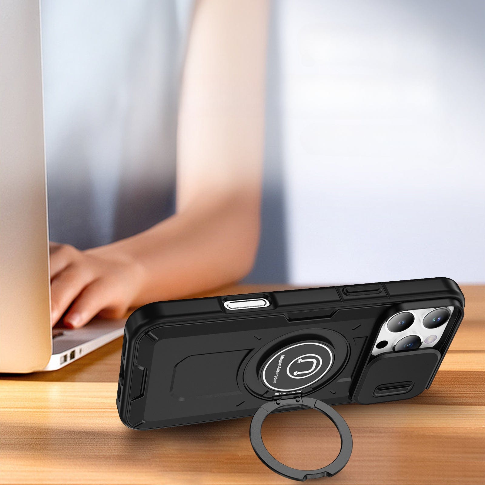 A person is using a laptop. In the foreground, an iPhone 16 Pro Max Rugged Magnetic Case with a sliding camera cover and shockproof protection rests on a wooden surface.