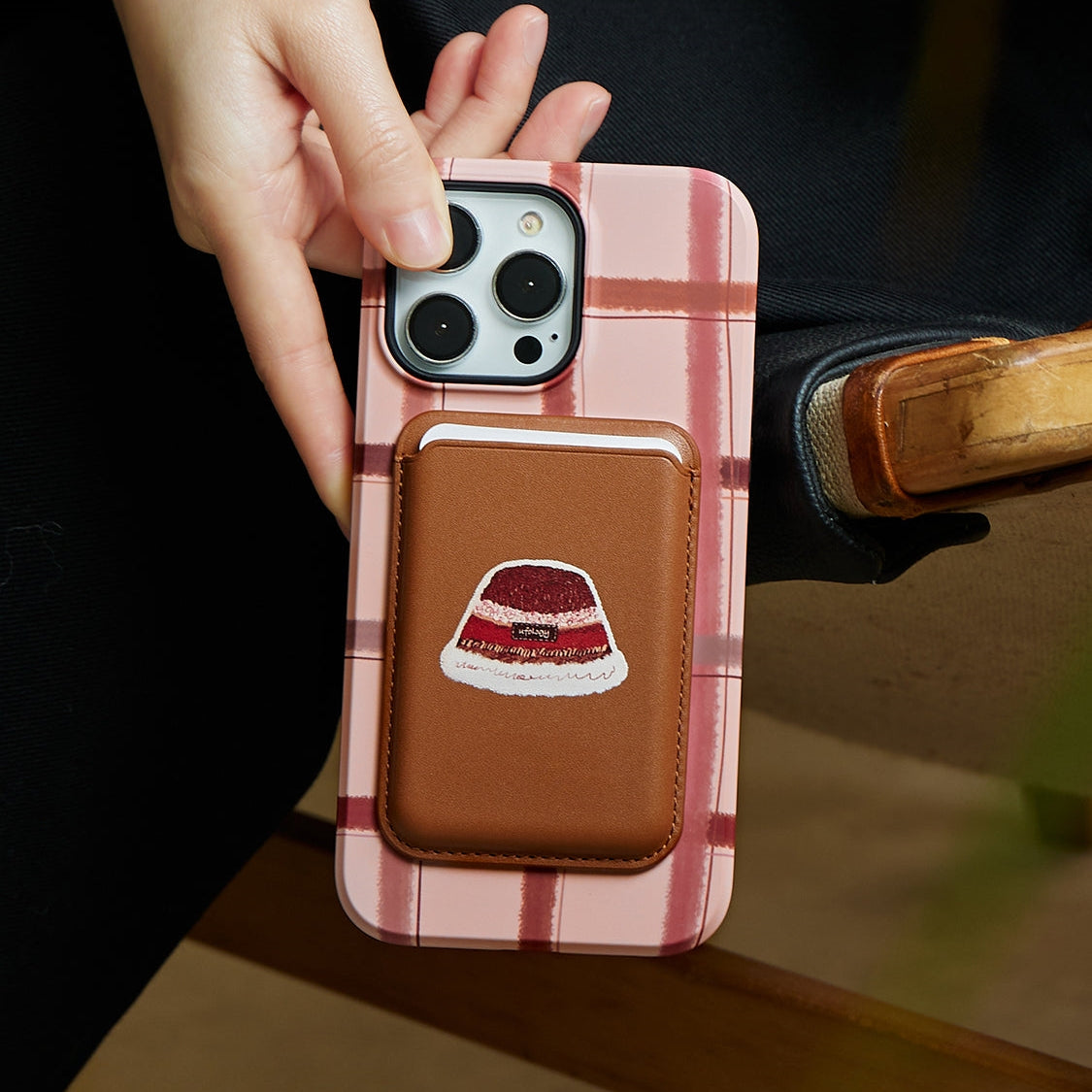 A hand holds a smartphone with a pink checkered case featuring a brown cardholder decorated with an embroidered dessert design.