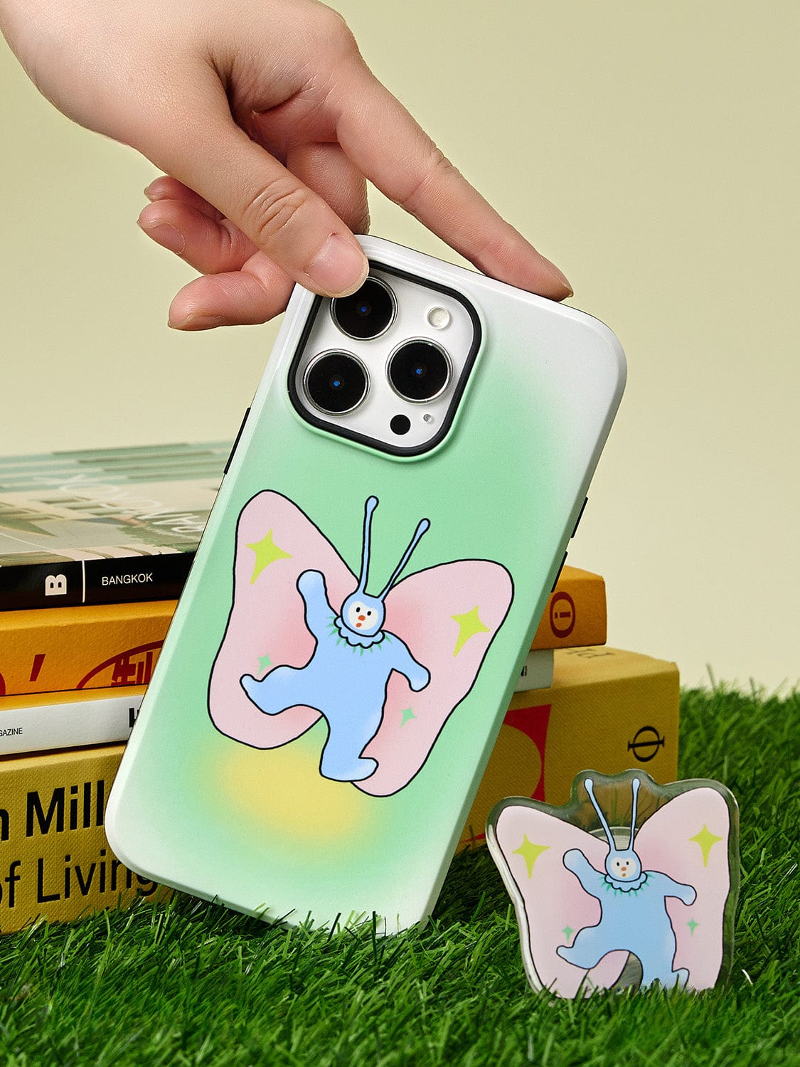 A hand holds the Golden Sunset MagSafe iPhone 16 Pro Max Case, showcasing its elegant gradient design and ring stand. The case features a cute butterfly illustration with a white face, blue body, pink wings adorned with yellow stars. The phone rests on a stack of books in a patch of grass, accompanied by a matching keychain beside it.