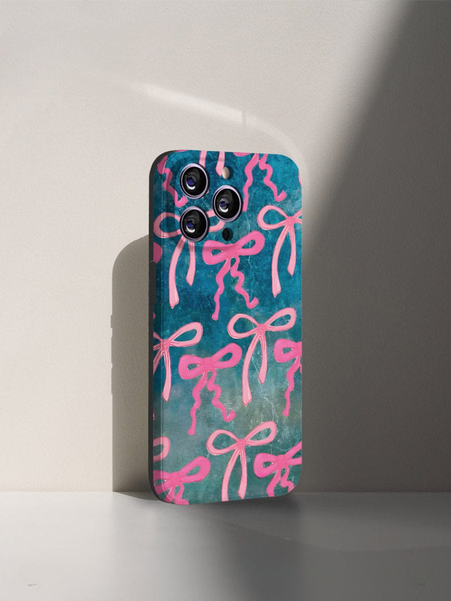 A smartphone fitted with the Pink Ribbon Charm iPhone 16 Pro Max Case, showcasing an artistic bow design with pink ribbons on a teal background, rests on a white surface. The case features precise cutouts for the camera lenses and is side-lit, casting a shadow on the wall.