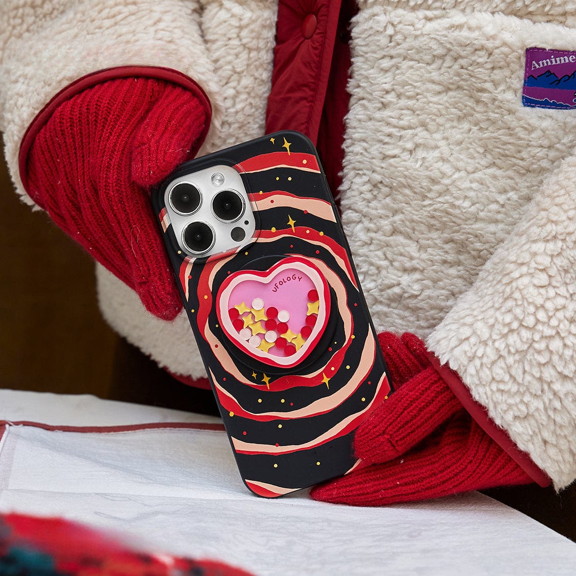 Close-up of a person with red gloves holding a Galaxy Heart MagSafe iPhone 16 Pro Max Case | Cosmic Design with Stand and an all-inclusive protective cover, featuring a heart-shaped cutout filled with small decorative elements and a name tag inside. The colorful case contrasts nicely against the fleece jacket they are wearing, which has a visible logo on the chest.