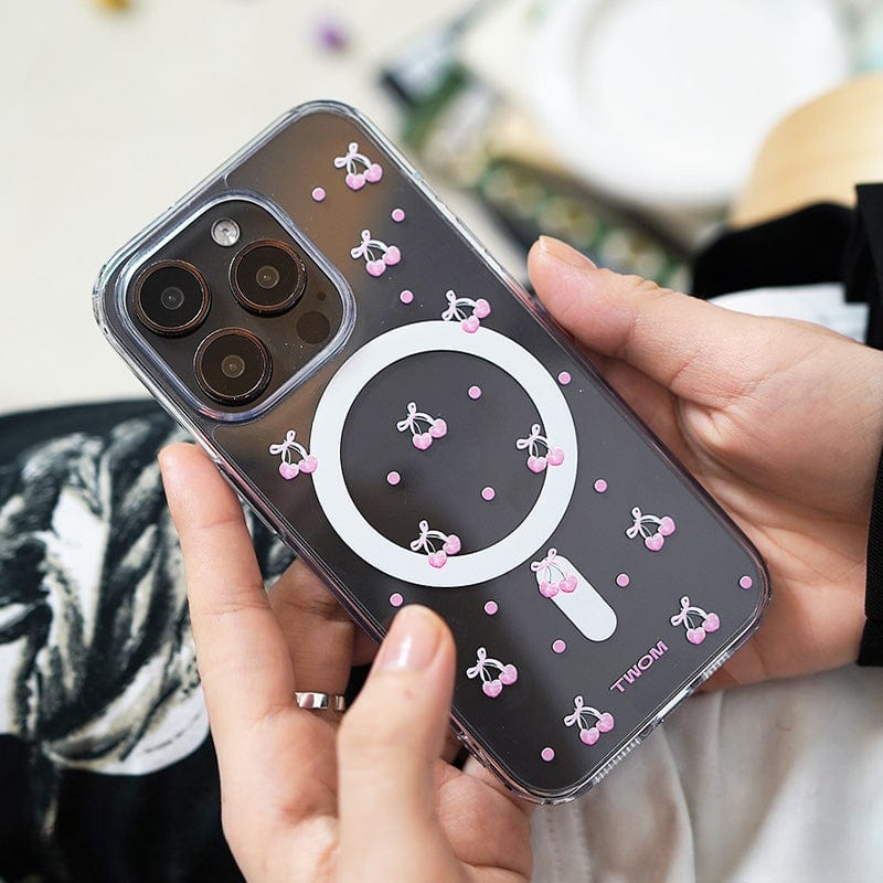 Close-up of a person holding the Cherry Blossom MagSafe iPhone 16 Pro Max Case. The case features a clear design with charming 3D pink cherry illustrations and showcases a visible circular magnetic circle. The person has light skin and is wearing black and white clothing.