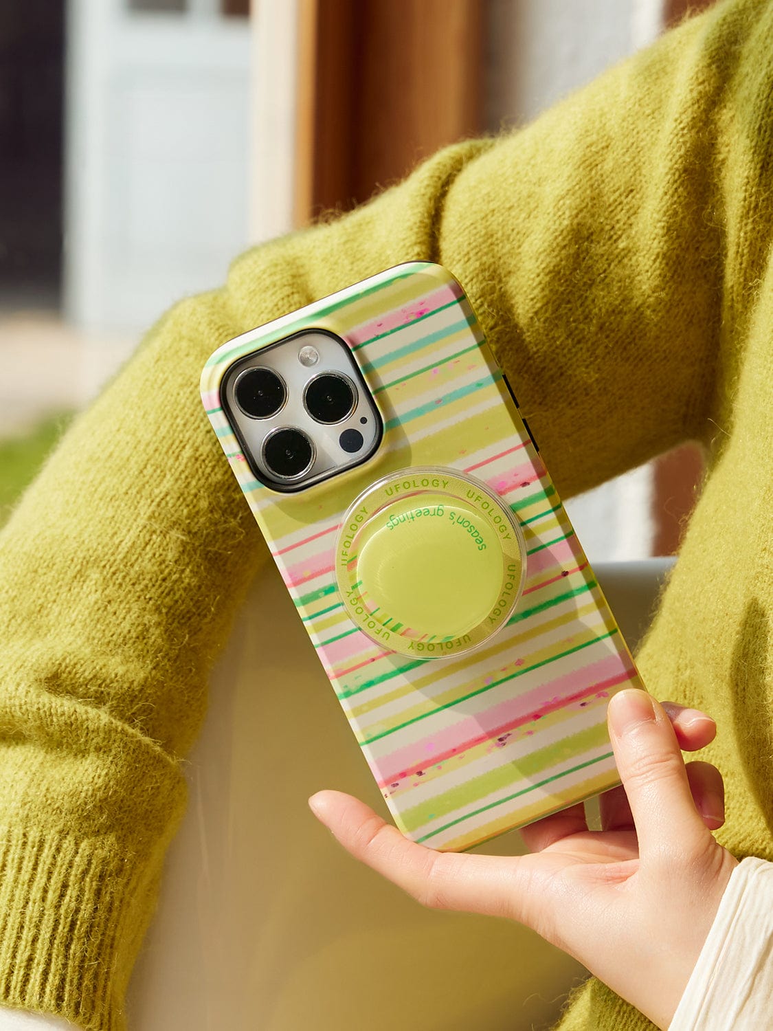 A person wearing a lime green sweater holds an iPhone 16 Pro Max with the Festive Stripes MagSafe Case, featuring cheerful holiday designs in green, pink, and yellow hues. The case also includes a lime green circular grip accessory attached to the back.