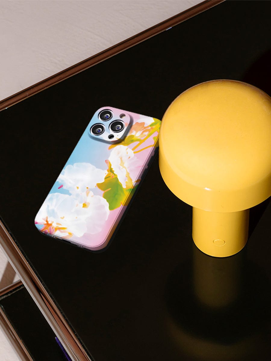 A Pastel Bloom iPhone 16 Pro Max Case, boasting a soft floral design and offering elegant all-inclusive protection, rests on a dark surface next to a yellow cylindrical lamp.