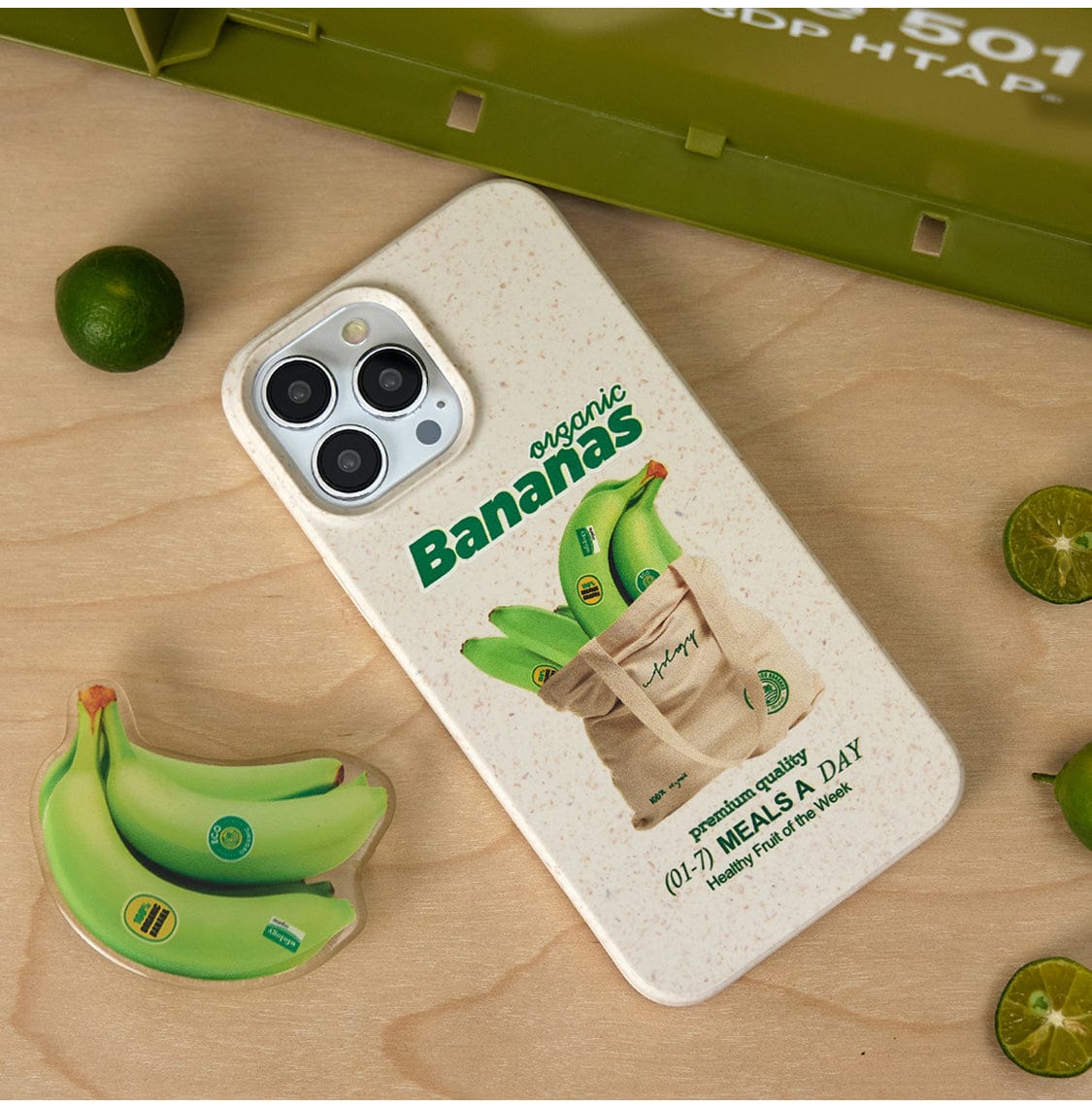 The Organic Bananas MagSafe iPhone 16 Pro Max Case features a delightful design of green bananas inside a beige tote bag. The text on the case reads "Organic Bananas, 0.17 MEALS A DAY, 'Healthiest Fruit of the Week.'" Placed on a wooden surface alongside a matching banana-themed pop socket and green limes, this all-inclusive protective cover exudes both charm and healthfulness.