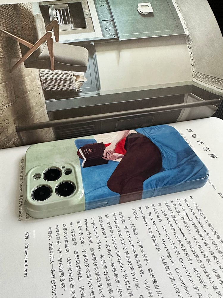 A magazine is open to a page displaying a modern interior room with a chair and a bookshelf. On top of the magazine lies the Relaxed Reader iPhone 16 Pro Max Case – Artistic and Protective, which features an image of a person reading a book while lying on blue fabric. The smartphone itself rests atop an open book filled with text.