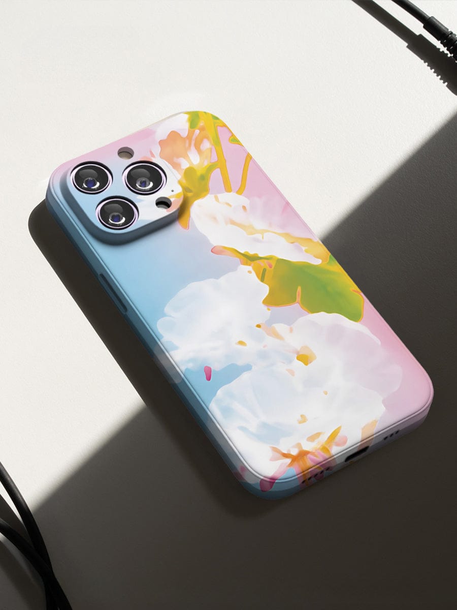 The Pastel Bloom iPhone 16 Pro Max Case, featuring a soft floral design, cradles the smartphone as it rests on a white surface, partially illuminated by sunlight. Wires are visible near the top and bottom edges of the image.