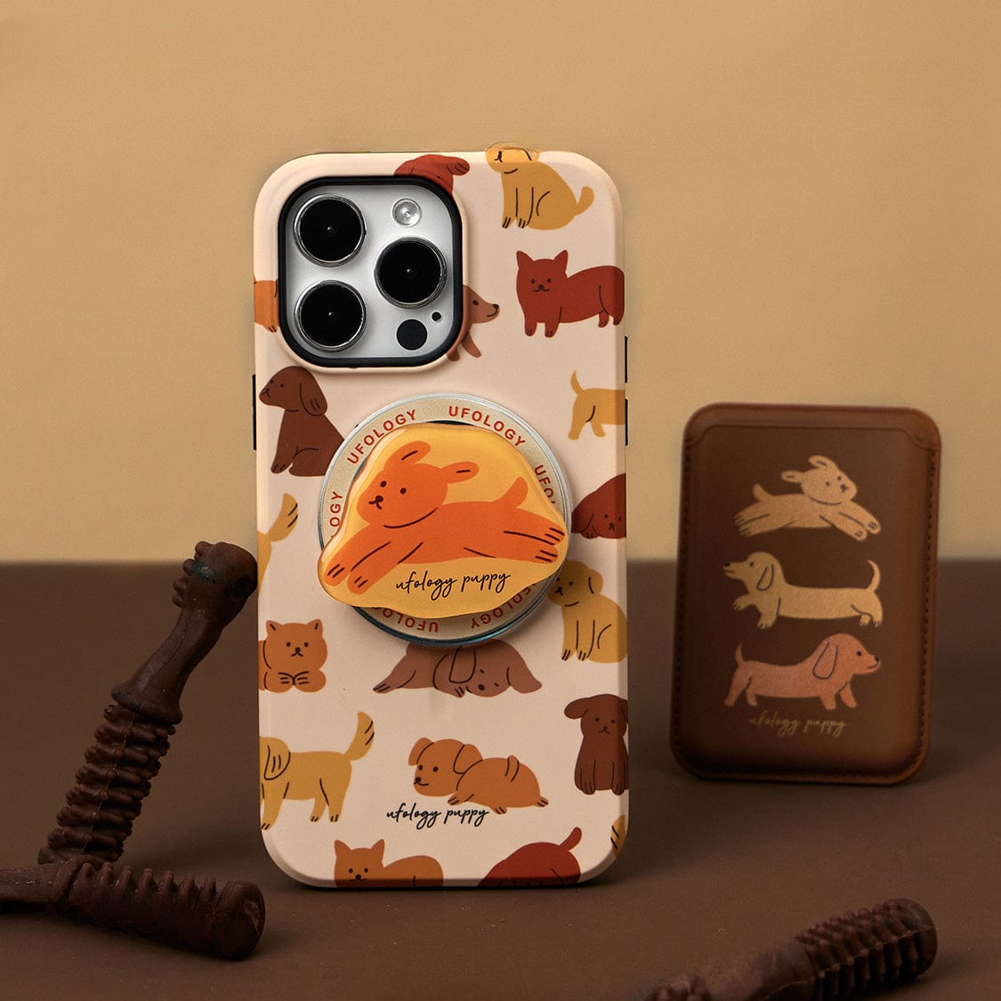 A smartphone with a Puppy Playtime MagSafe iPhone 16 Pro Max Case, featuring a cute dog design and integrated stand, is positioned upright near a brown wallet adorned with a dachshund pattern. The background showcases a neutral brown and beige color scheme.