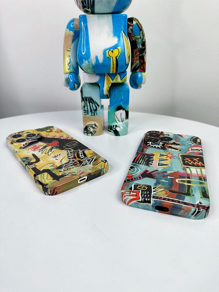 A photo of two vibrant Urban Graffiti iPhone 16 Pro Max cases from the Bold Street Art Collection with abstract patterns lying on a white surface in front of a standing figurine. The figurine, featuring a similar abstract design, stands upright in the background.