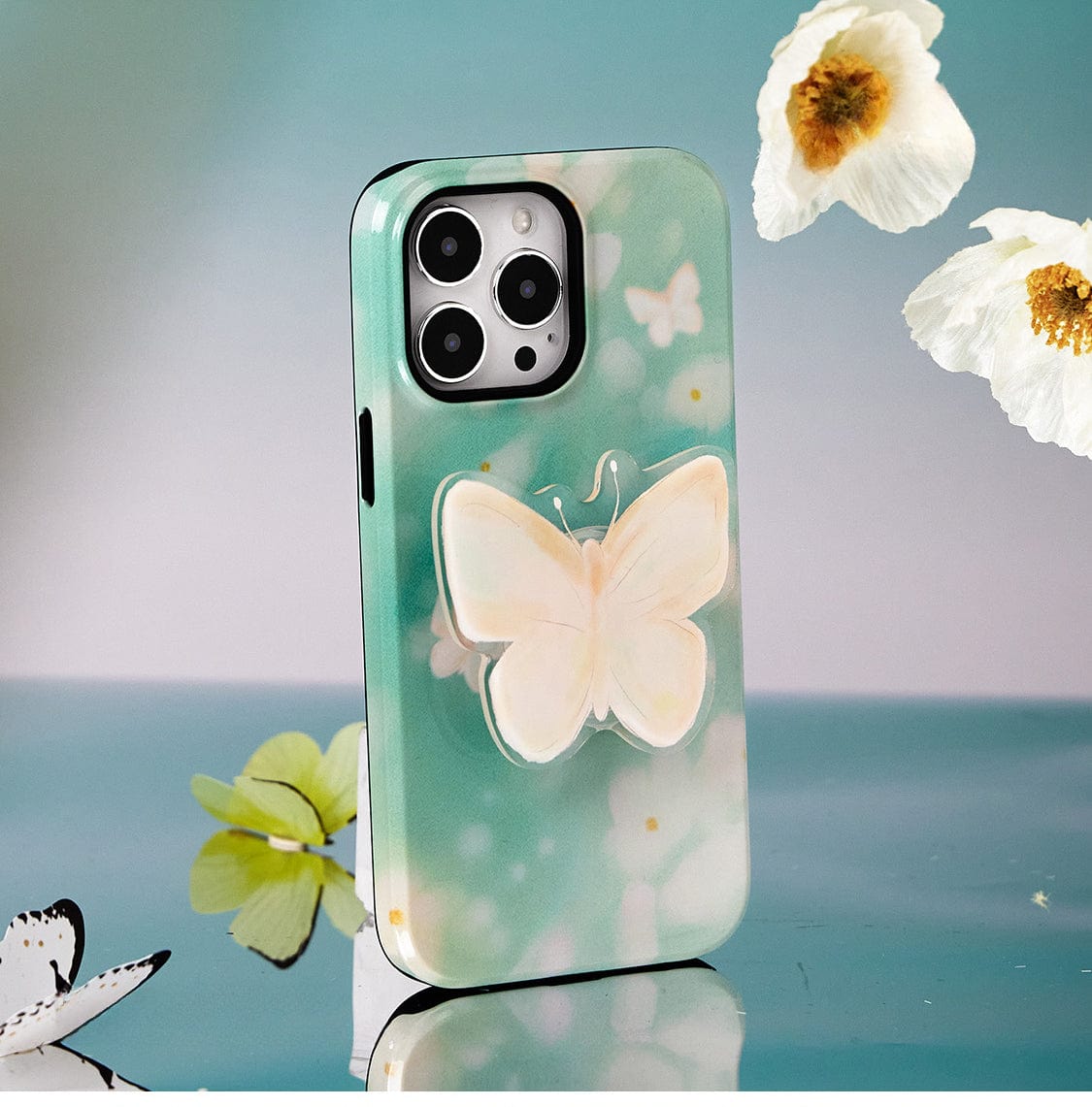 The Butterfly Blossom MagSafe iPhone 16 Pro Max Case, featuring a white butterfly design and an attached butterfly ornament, is displayed upright against a backdrop of flowers and butterflies.