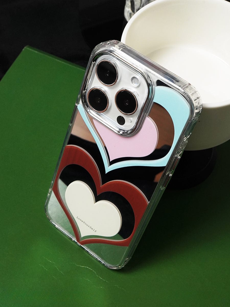 A Retro Heart Layers iPhone 16 Pro Max Case with a Vibrant Love Yourself Design, showcasing multicolored hearts, is displayed on a green surface, partially set against a white bowl.