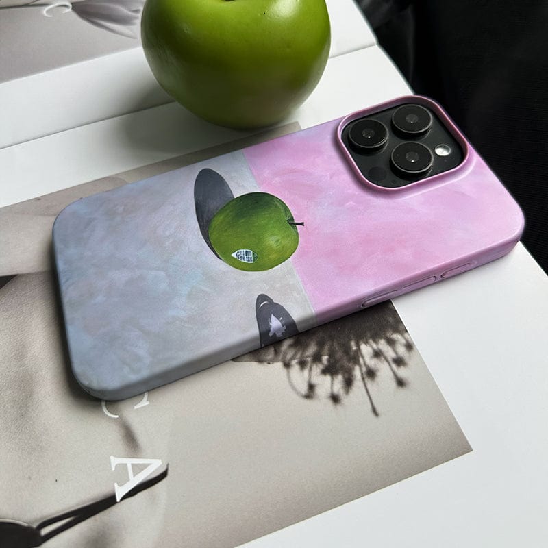 A smartphone in the Minimalist Green Apple iPhone 16 Pro Max Case, which showcases an artistic still life design featuring a green apple partially obscured by a floating black hat. The background of the case smoothly transitions from gray to pink. The phone is placed on an open book, with a real green apple close by.