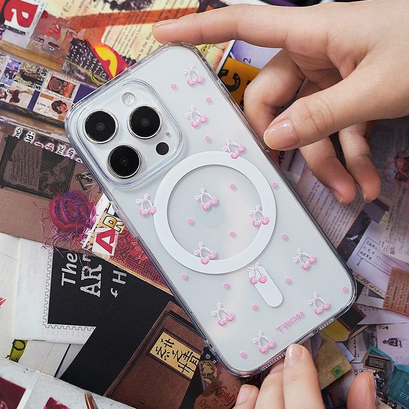 A person holds a Cherry Blossom MagSafe iPhone 16 Pro Max Case, distinguished by its cute 3D cherry design and all-inclusive protective cover. The case features small pink cherries and a white ring-shaped phone holder in the center. The background showcases an array of colorful magazine clippings and scattered envelopes.