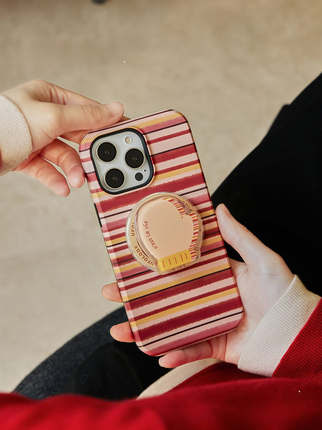 A hand holds an Autumn Stripes MagSafe iPhone 16 Pro Max Case, featuring colorful horizontal red, pink, and beige stripes. The case showcases a peach-colored, heart-shaped phone grip attached to the back. The background is a blurred indoor setting.