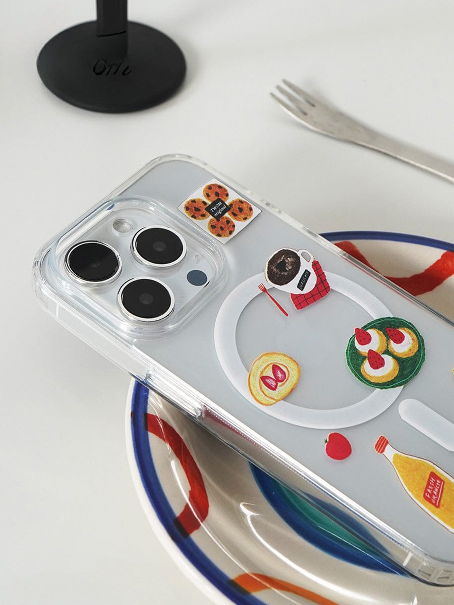 A smartphone with a Brunch Time MagSafe iPhone 16 Pro Max Case, adorned with cute 3D food design stickers of pancakes, jam, avocado toast, coffee, and utensils, rests on a multicolored plate. A nearby metal fork and part of a black object are visible in the background.