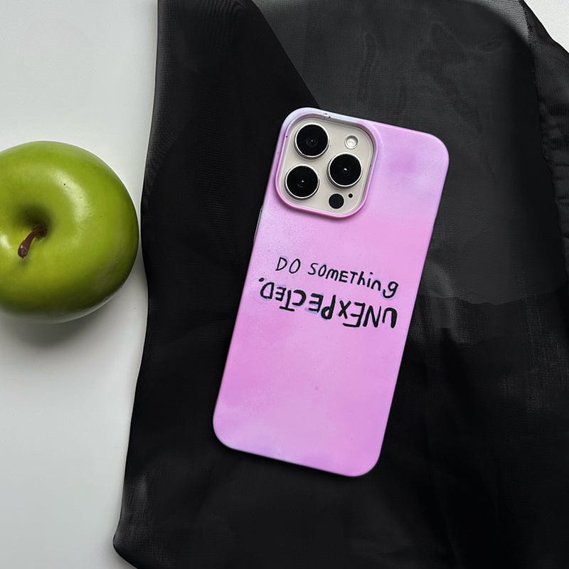 An iPhone 16 Pro Max with an Inspirational Pink case featuring the quote "Do Something Unexpected" printed in an inverted orientation lies on a black fabric. Next to the phone is a green apple.