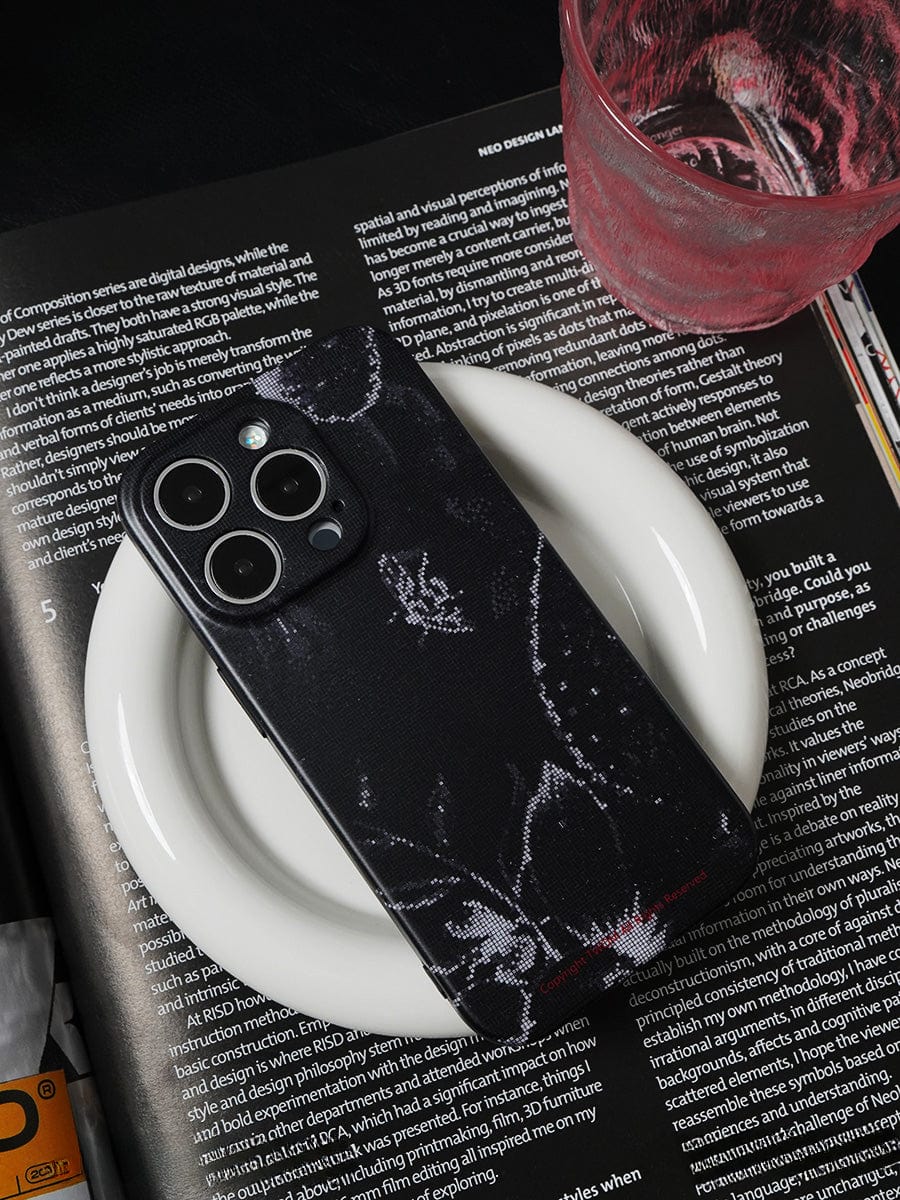 A smartphone with a Noir Floral iPhone 16 Pro Max Case, featuring an elegant black and white design, rests on a white plate atop an open book or magazine. A transparent glass with a pink rim containing liquid is placed nearby on the open pages. The background is primarily dark, highlighting the objects.