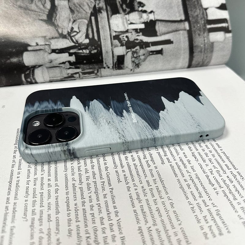 A close-up of a smartphone adorned with a Monochrome Waves iPhone 16 Pro Max case featuring a minimalist design and the quote "Better Late Than Never," laying on top of an open book. In the background, part of another open book with an old black-and-white photograph is visible. The phone is placed screen-down, showcasing the back and camera lenses.