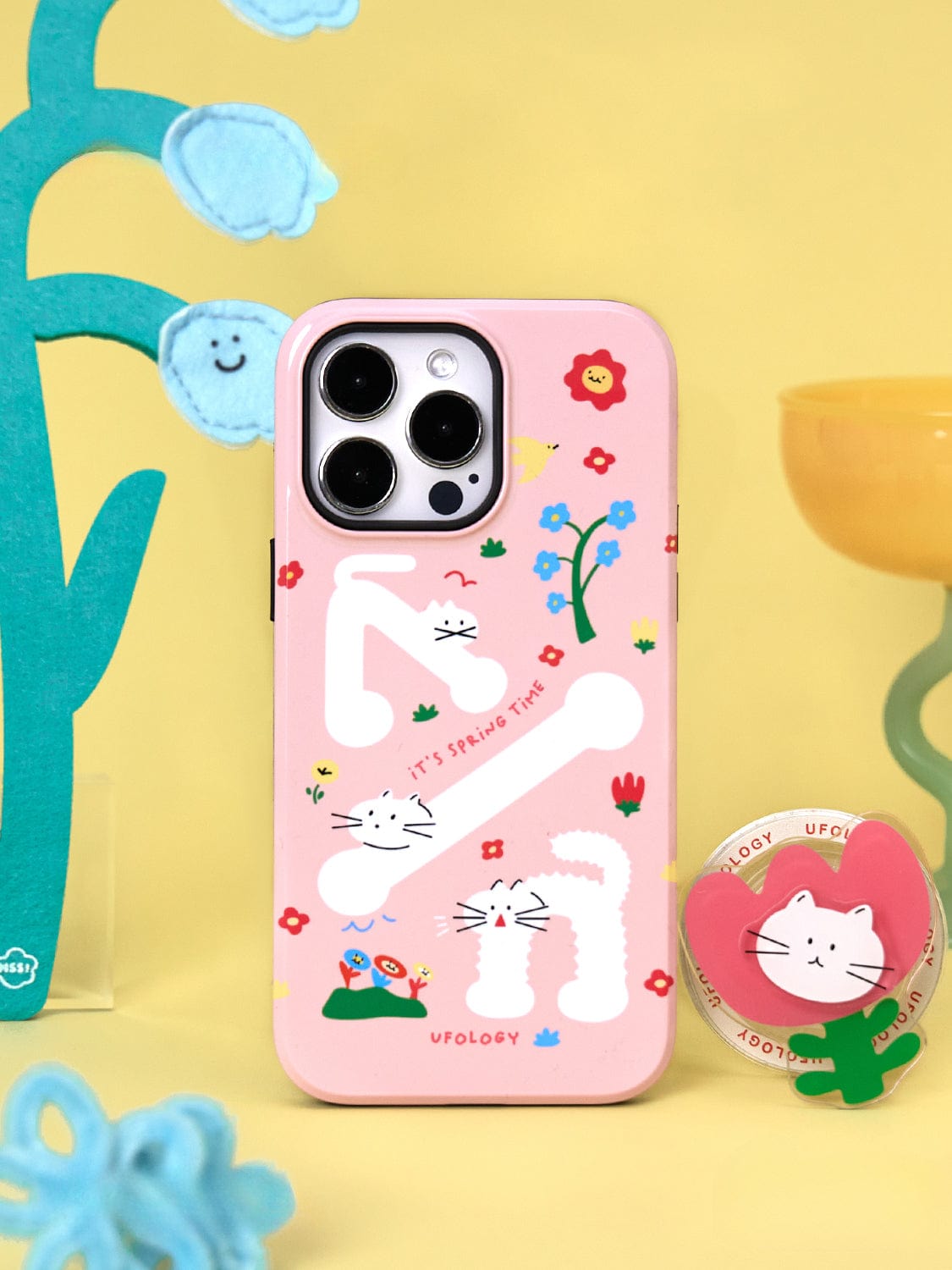 A smartphone with a Spring Kitty MagSafe iPhone 16 Pro Max case, featuring white cartoon cats and playful floral design, complete with a cat stand, placed on a yellow surface surrounded by colorful decorative items.