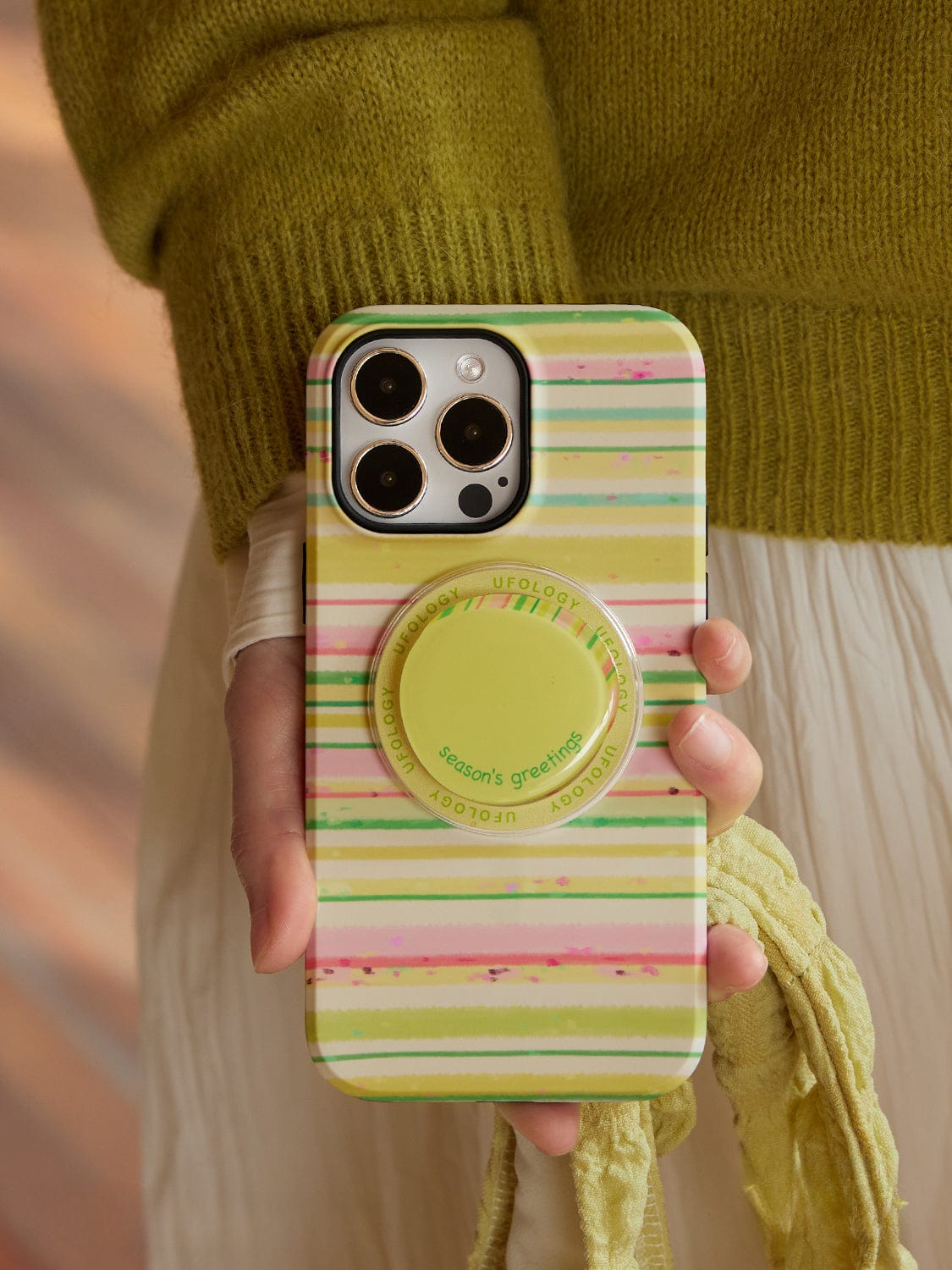 A person holds an iPhone 16 Pro Max enclosed in a Festive Stripes MagSafe Case, featuring a colorful striped design in shades of green, pink, and cream. The case includes a round pop-out grip. The person is dressed in a green sweater and white skirt, with a yellow item draped over their hand.