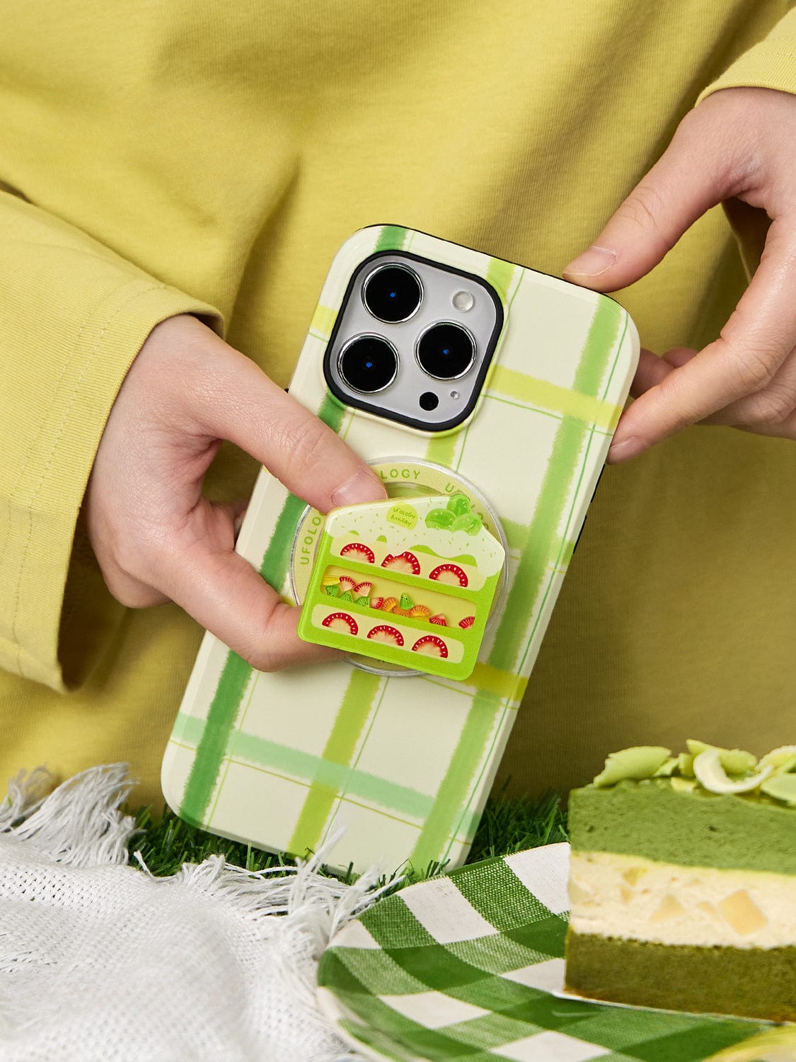 A person holding the Fresh Picnic MagSafe iPhone 16 Pro Max Case featuring a green plaid design and a sandwich stand, next to a slice of matcha cake on a white cloth.