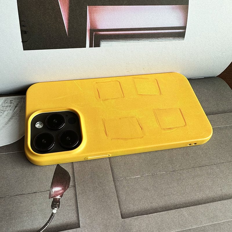 A Bold Yellow Textured iPhone 16 Pro Max Case, featuring a modern art-inspired minimalist design and durable protection with camera cutouts, lies on an open magazine. The case shows visible wear and impressions from being previously folded, while the magazine page it rests on features geometric shapes and a light fixture.