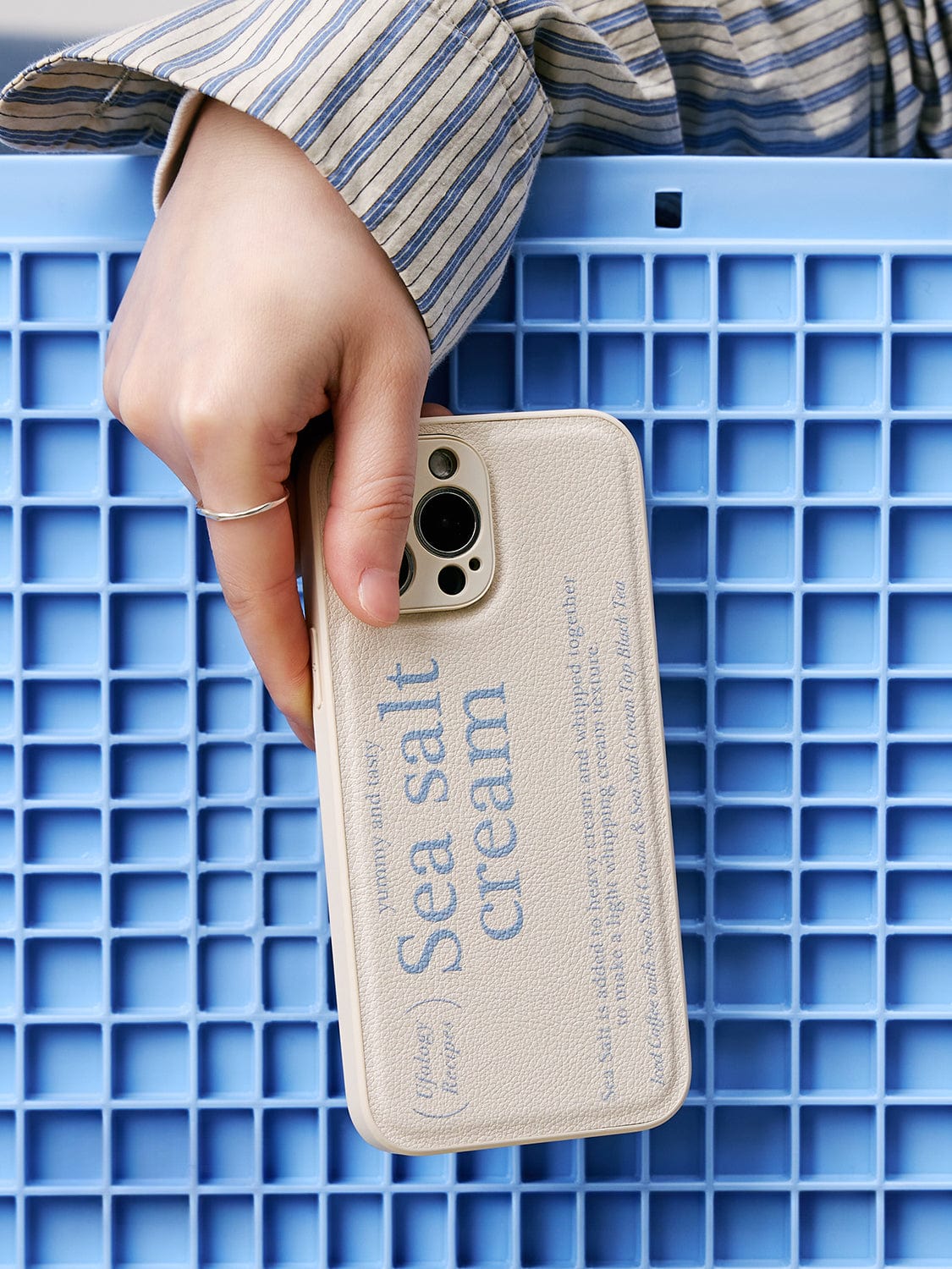 A hand holding an iPhone 16 Pro Max encased in a Sea Salt Cream MagSafe Case, featuring a minimalist textured design with the text "Sea salt cream" printed on it, set against a textured blue background.