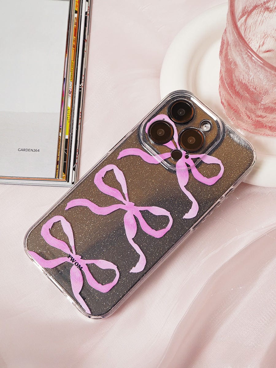 A smartphone encased in a Pink Ribbon Bow Transparent iPhone 16 Pro Max Case, featuring a sparkling clear design and elegant protective cover, rests on a light pink fabric. The phone sits adjacent to a clear glass with a pink hue, and an open book or magazine is partially visible in the background.