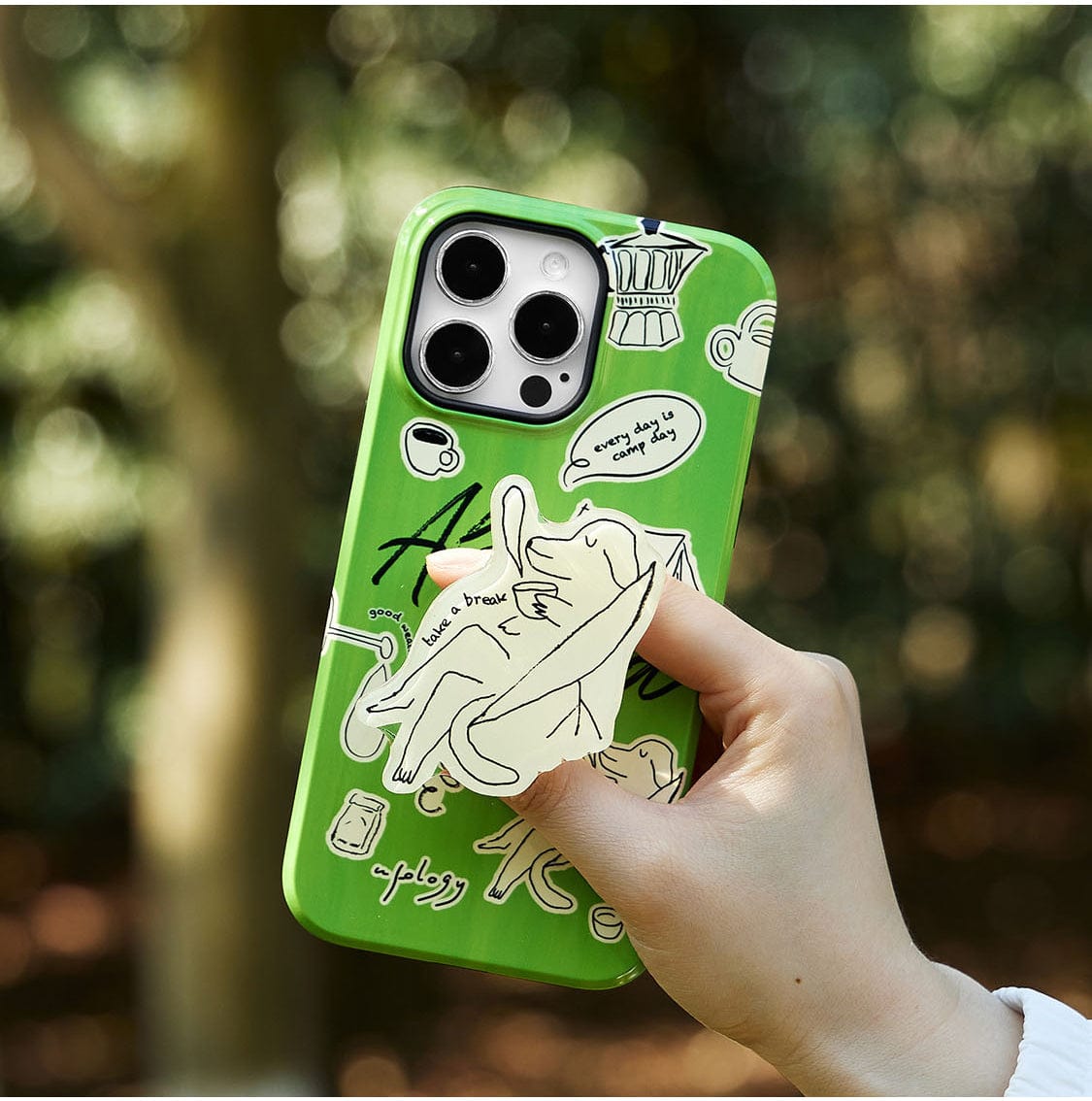 A hand holding a Lazy Day Elephant MagSafe iPhone 16 Pro Max Case with a playful green background featuring sketches of a toga-clad figure and various doodles, including text phrases like "take a break" and "apply tones." The background shows blurred greenery.