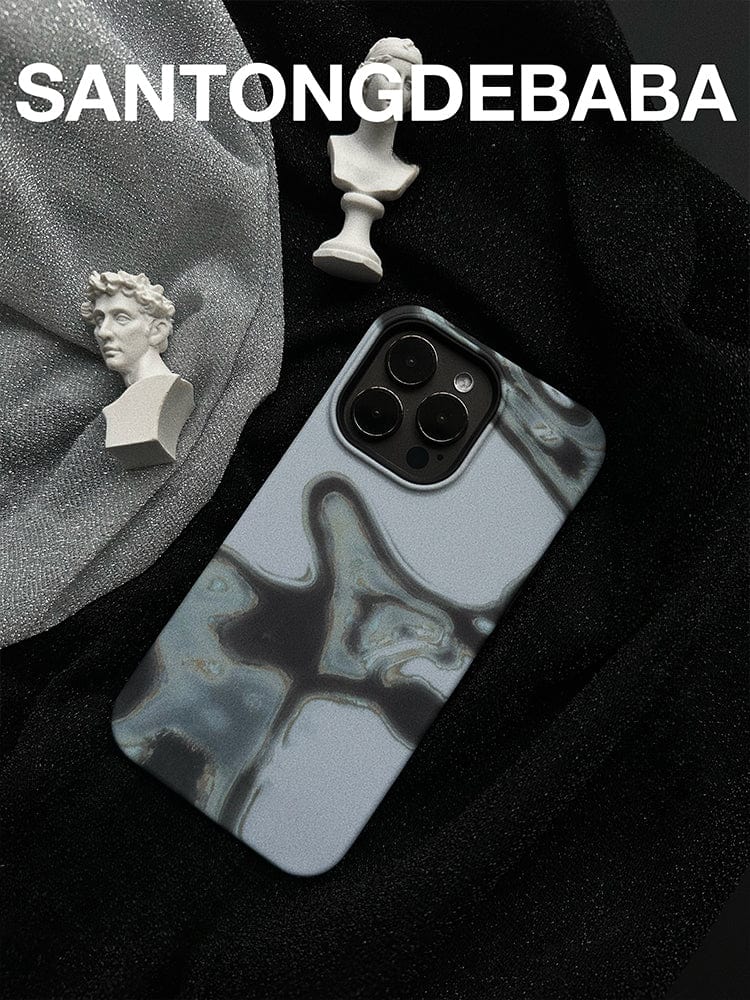 An Abstract Metallic Flow iPhone 16 Pro Max case, featuring a sleek modern art design with gray and black patterns, is placed on a dark fabric background. Next to it are two small white sculptures: one of a bust and the other of a vase. The text "SANTONGDEBABA" is written at the top.