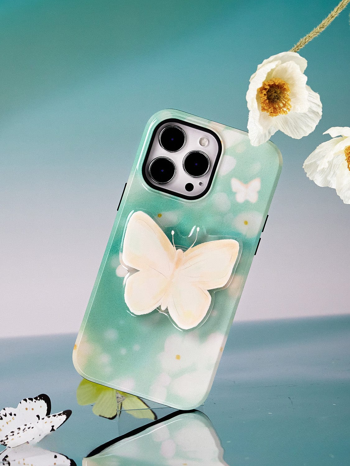 The Butterfly Blossom MagSafe iPhone 16 Pro Max Case, featuring elegant butterfly designs on a teal background and displayed upright near white butterfly decorations and flowers, is the perfect accessory for your iPhone 16 Pro Max.