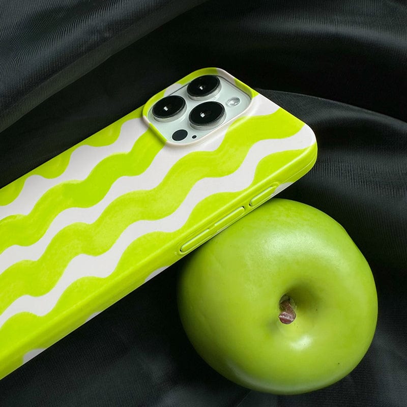 A smartphone with a Vibrant Wavy Line iPhone 16 Pro Max Case - Bold Lime Green & Cream Design, Ultra-Slim, Protective Cover is placed on a dark fabric background, next to a green apple. The phone's camera is visible, and the apple is positioned to the right of the device.