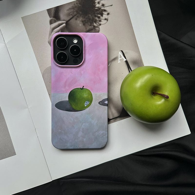 A phone with a Minimalist Green Apple iPhone 16 Pro Max Case - Artistic Still Life Design with Full Protection, featuring a pink and gray aesthetic and a green apple illustration, rests on an open black and white magazine. Next to the phone lies a real green apple. The scene is set against a dark background, highlighting the contrast between the bright green apple and surrounding elements.