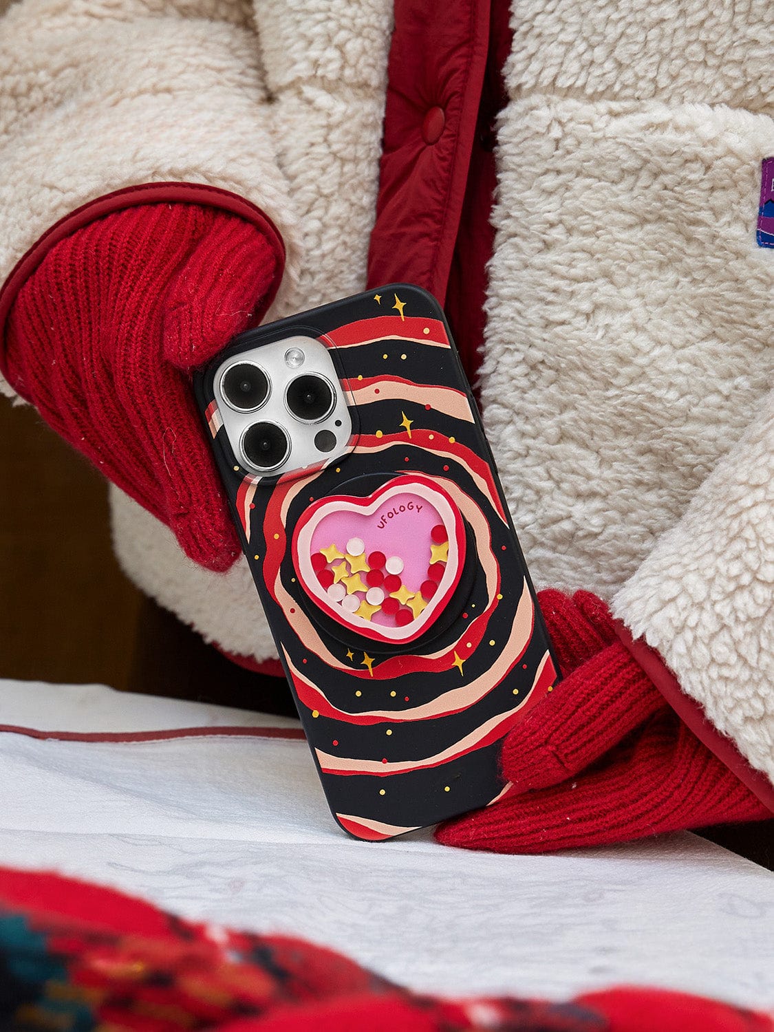 A person wearing a white fleece jacket and red gloves holds an iPhone 16 Pro Max with a Galaxy Heart MagSafe Case. The case boasts a colorful, cosmic design and features a heart-shaped compartment filled with small yellow and white objects, with the word "Jadore" inscribed inside the heart.