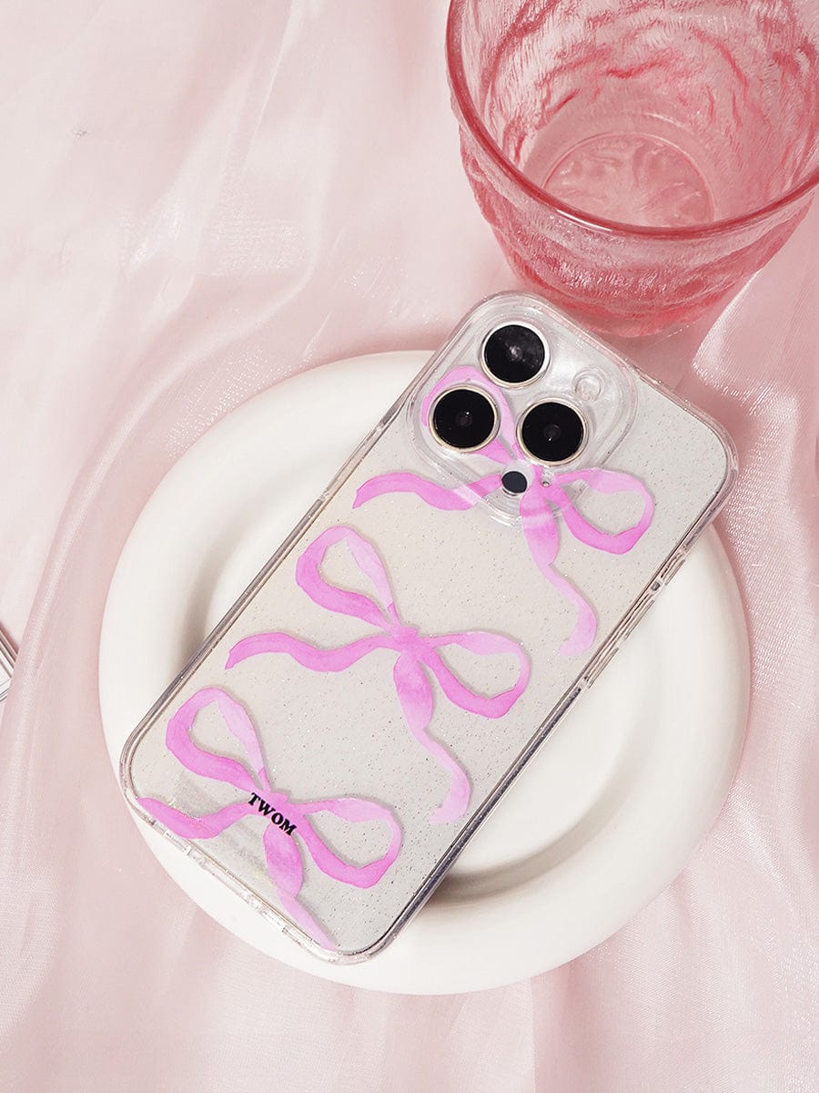 The Pink Ribbon Bow Transparent iPhone 16 Pro Max Case, featuring a sparkling clear design and elegant all-inclusive protective cover, is placed on a white plate. Next to it is a pink textured glass on a soft pink fabric background.