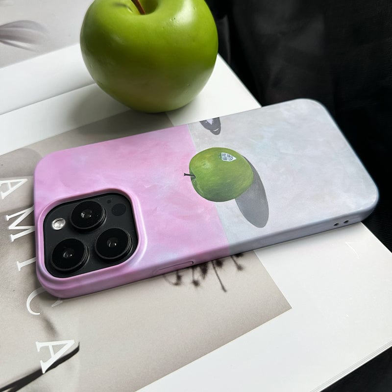 A smartphone with the Minimalist Green Apple iPhone 16 Pro Max Case, showcasing an artistic still life design of a green apple floating in a surreal landscape with shadows beneath it. An actual green apple is placed nearby on a white surface, with a book partially visible in the background.