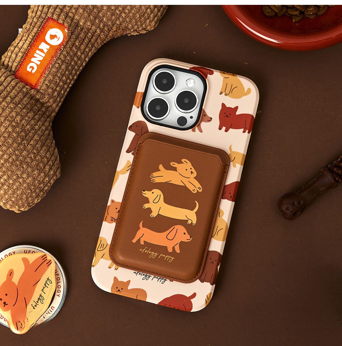 A Puppy Playtime MagSafe iPhone 16 Pro Max Case featuring a cute dog design and a stand, along with a brown card holder, is placed alongside a bone-shaped dog toy, a round dog-themed item, and a bowl of kibble.