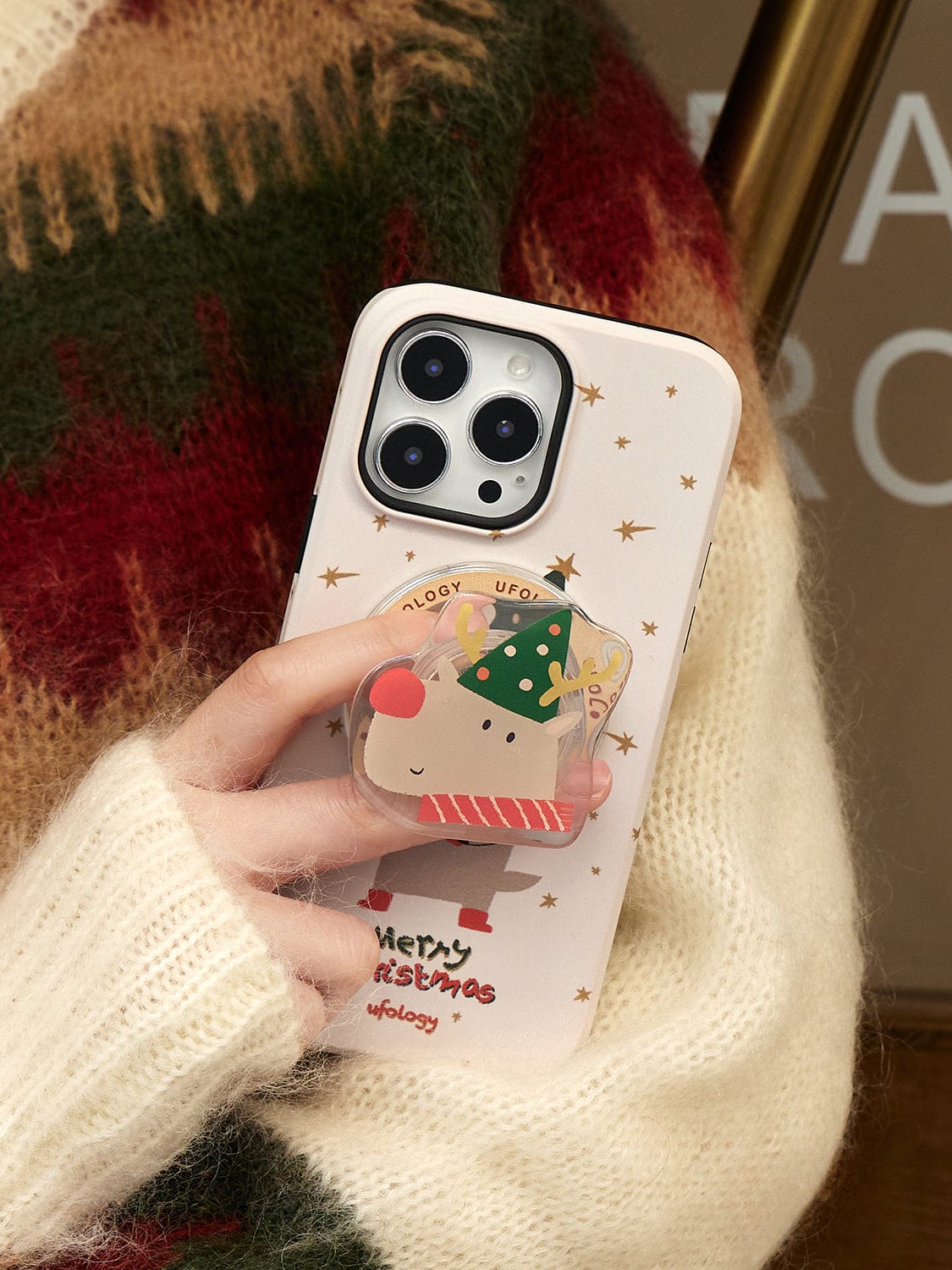 A hand holding an iPhone 16 Pro Max with a Rudolph Joy MagSafe iPhone 16 Pro Max Case featuring a festive reindeer design and "Merry Christmas" text. The person is wearing a cozy, multicolored sweater.

