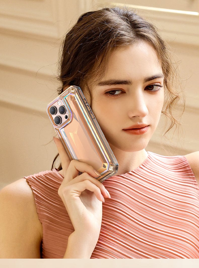 A woman with light brown hair holds a Shockproof 360° Clear Anti-Fall Protective Cover with a 3D Gem Design for an iPhone 16 Pro Max to her ear, wearing a sleeveless, pleated, light pink top. The background appears to be an indoor setting with neutral-toned walls.