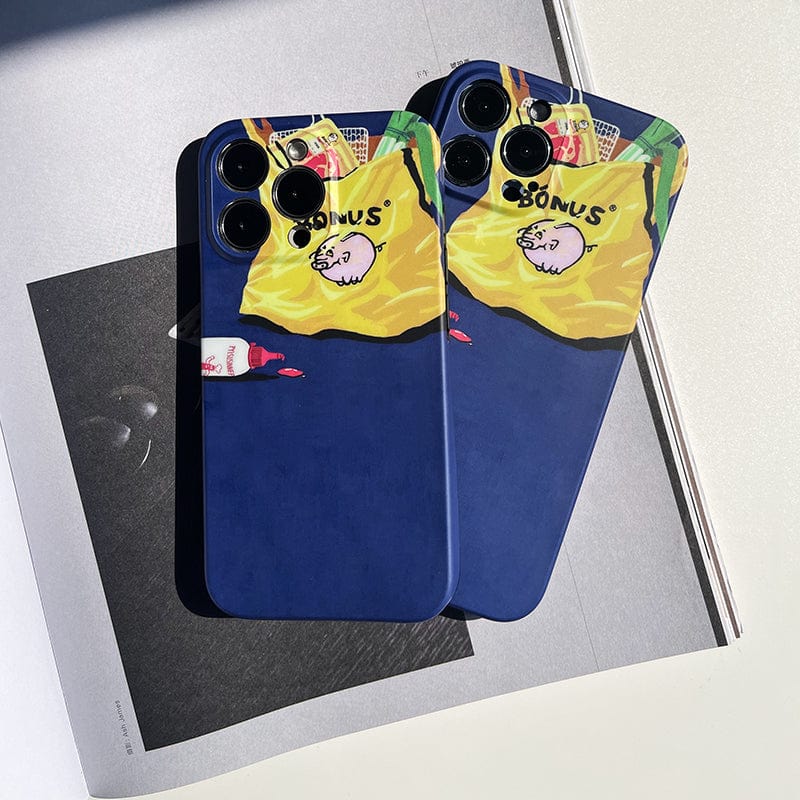 Two iPhone 16 Pro Max devices with colorful Quirky Grocery Bag cases featuring a whimsical design are placed side by side on an open book. The cases display a yellow and blue shopping bag illustration with a cartoon pig and the text "BONUS." Nearby is a red and white object that resembles a marker.