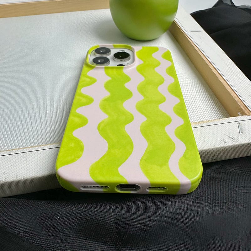 A close-up of the Vibrant Wavy Line iPhone 16 Pro Max Case - Bold Lime Green & Cream Design, Ultra-Slim, Protective Cover showcases its striking wavy pattern. The phone is placed on a white fabric surface, with a green apple partially visible in the background. The bottom part of the phone, including ports and speakers, is clearly visible.