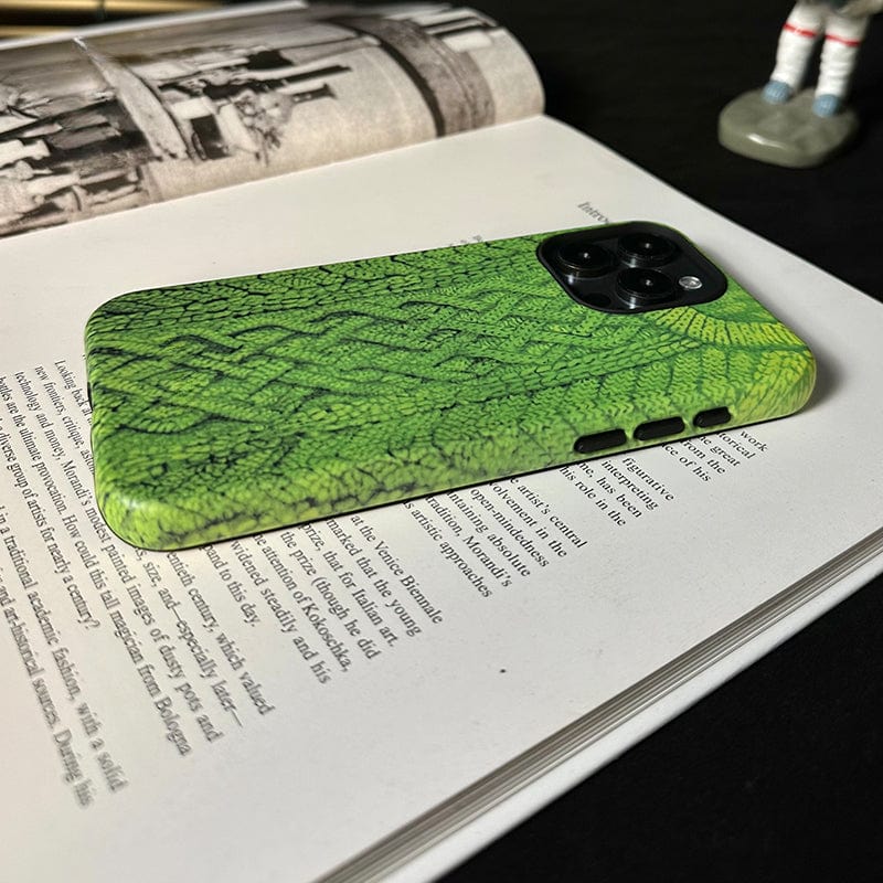 A smartphone with a Cozy Knitted Sweater iPhone 16 Pro Max Case - Textured Green Design for Warmth and Protection, resembling reptile skin, is placed on an open book. In the background, a figurine stands on a surface. The phone's camera module is visible, and the case has precise cutouts for buttons.