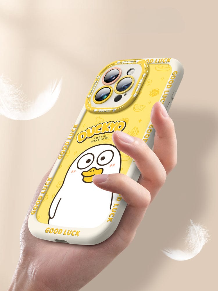 A hand holding an iPhone 16 Pro Max with the Yellow Duck Cartoon Case - a soft liquid silicone protective cover featuring a cute cartoon duck and the text "DUCKYO." The case, which is shockproof and anti-yellowing, has white edges decorated with the words "GOOD LUCK" repeating around the borders.