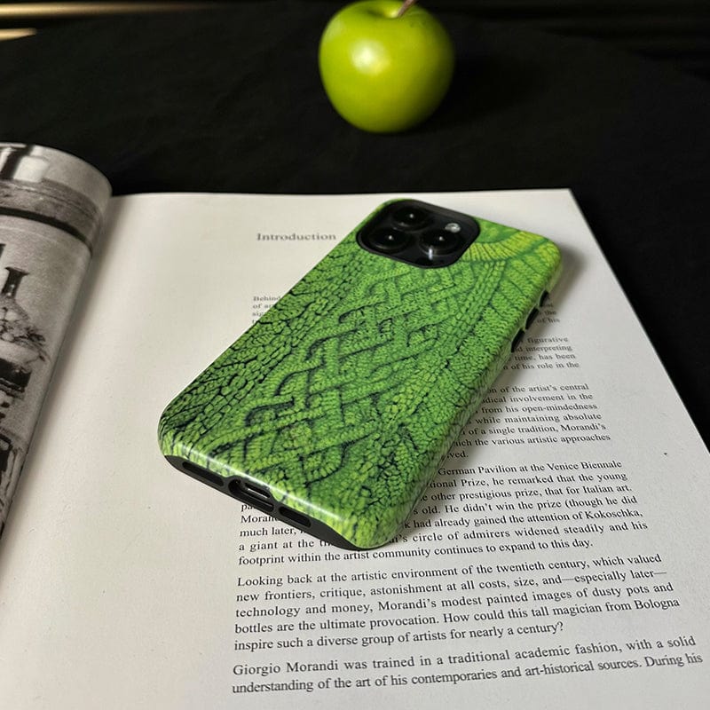 A smartphone with a Cozy Knitted Sweater iPhone 16 Pro Max case, green and textured for warmth and protection, rests face down on an open book. An apple sits in the background on the dark surface. The book is opened to a page displaying text in English and a blurred black-and-white image.