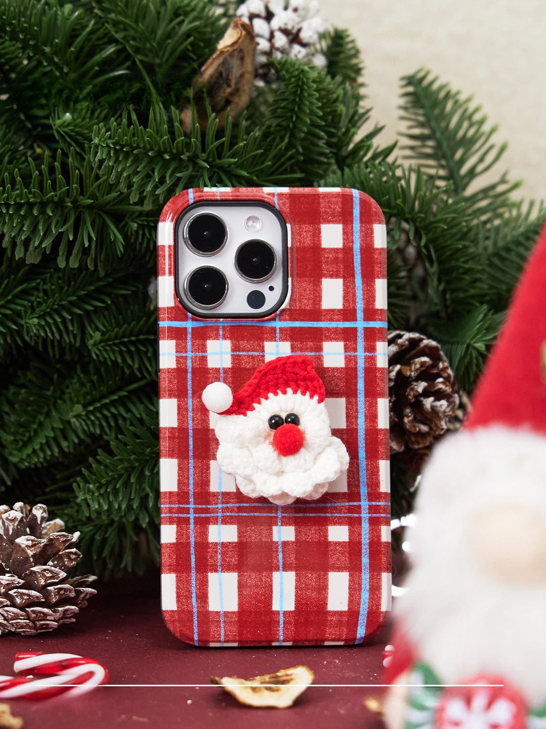 A Festive Knitted MagSafe iPhone 16 Pro Max case with a holiday plaid design featuring Santa and tree stands rests against a backdrop of green pine branches, pinecones, and holiday decorations.