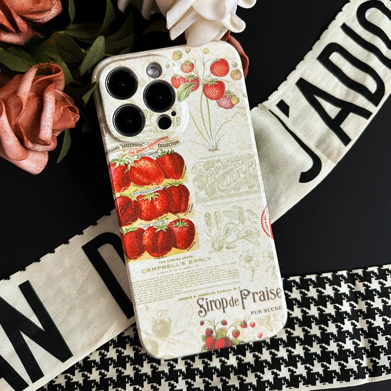 The Vintage Strawberry Print iPhone 16 Pro Max Case - Elegant and Protective showcases vintage botanical illustrations of tomatoes and strawberries surrounded by script text. The phone case is set on a black surface near a piece of white fabric with black text and pink flowers.