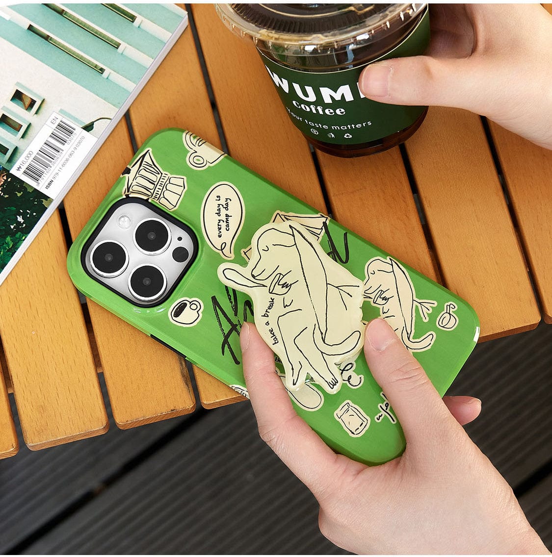 Hands holding a smartphone in a Lazy Day Elephant MagSafe iPhone 16 Pro Max Case with a playful green design. An iced coffee cup and a book are on a wooden table.