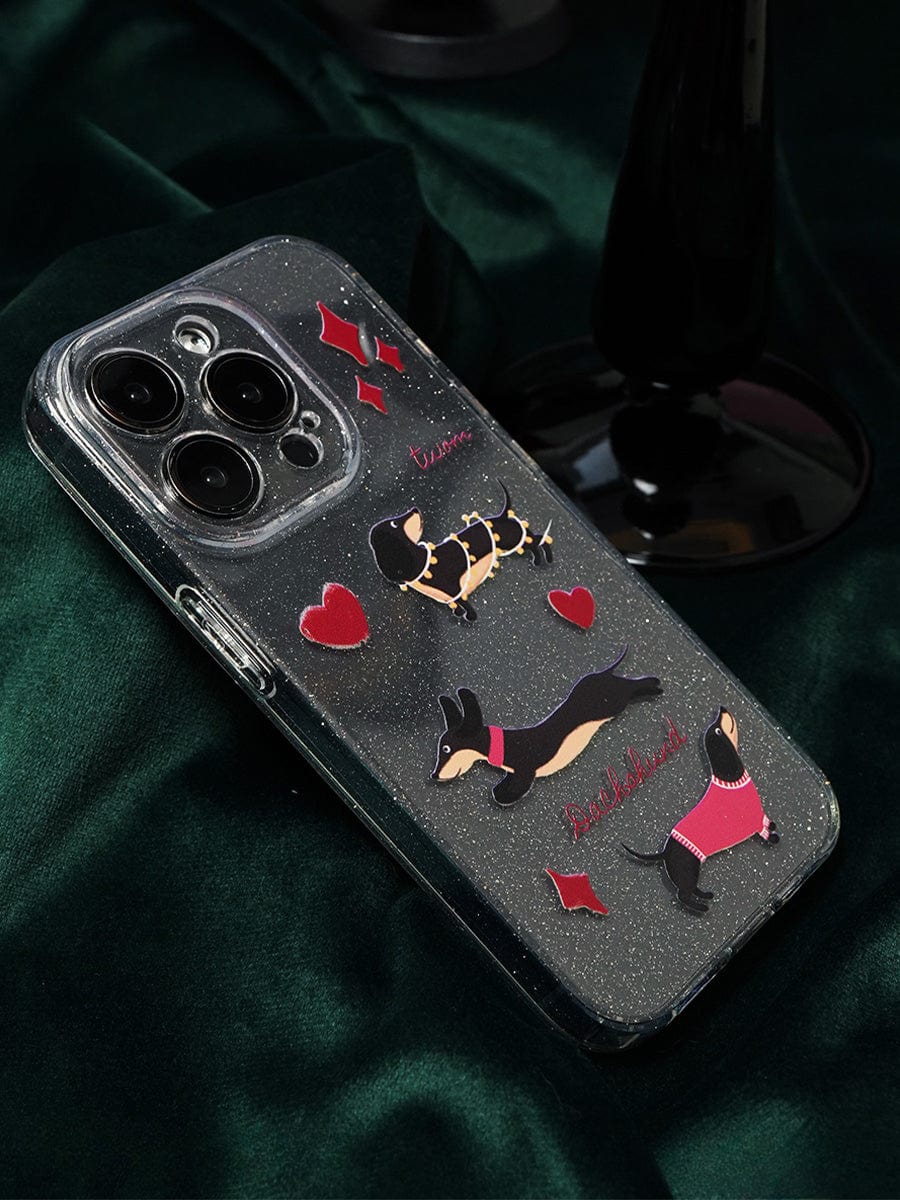 A smartphone encased in a clear sparkle "Dachshund Love iPhone 16 Pro Max Case" featuring cartoon dachshunds in sweaters, illustrated bones, and red hearts. The phone rests on a dark green fabric background with part of a black object visible in the top-right.