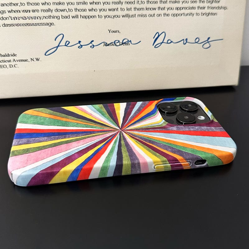 A smartphone with a Vibrant Burst iPhone 16 Pro Max Case in a Colorful Radiant Stripes Design lies on a black surface. Behind it, there is part of a white box displaying a message and a handwritten signature in blue ink.